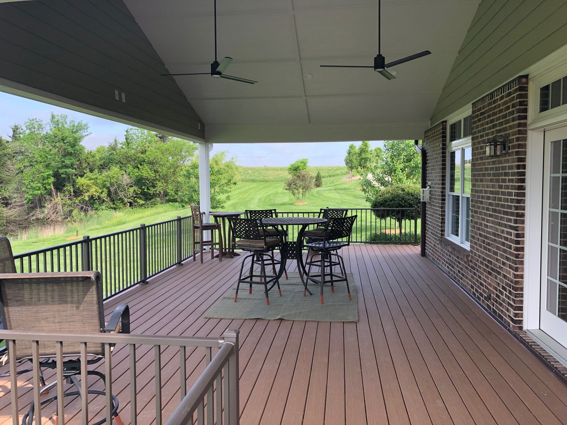 A covered outdoor deck with a table, chairs, and a view of a green field. The deck is brown with black railings.