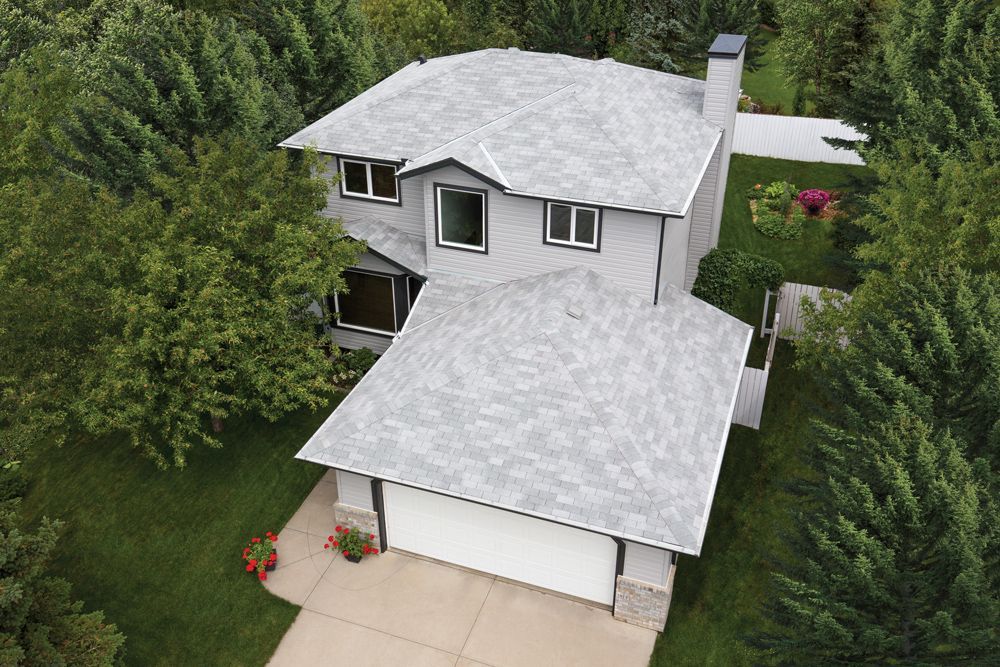 Aerial view of a two-story house with a light gray roof and siding, surrounded by green trees and grass.