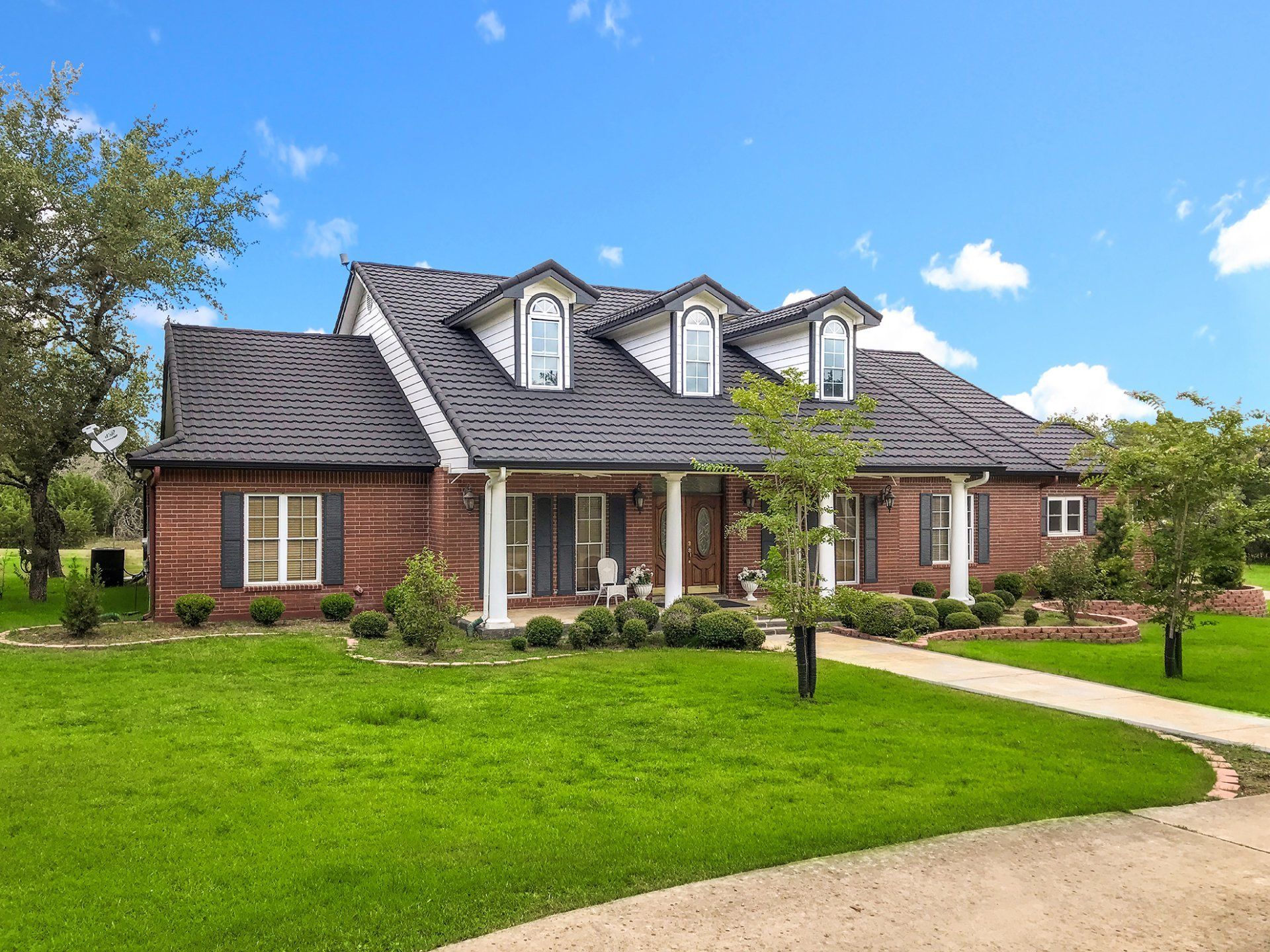 Brick house with a dark roof, white columns, and dormer windows under a blue sky. Lush green lawn in front.