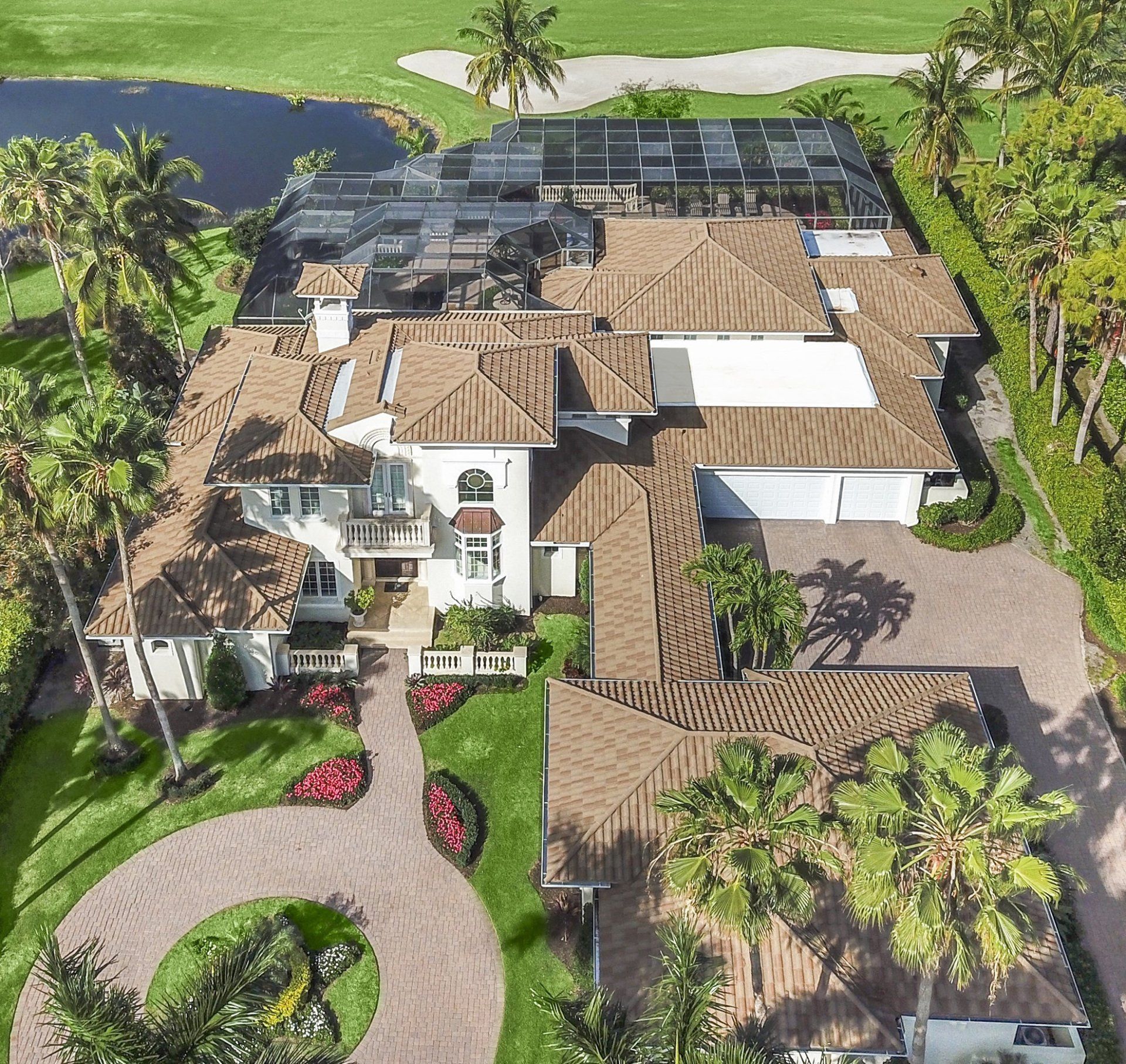 Aerial view of a large white house with a brown tile roof, surrounded by green lawn and palm trees, next to a golf course and a pond.