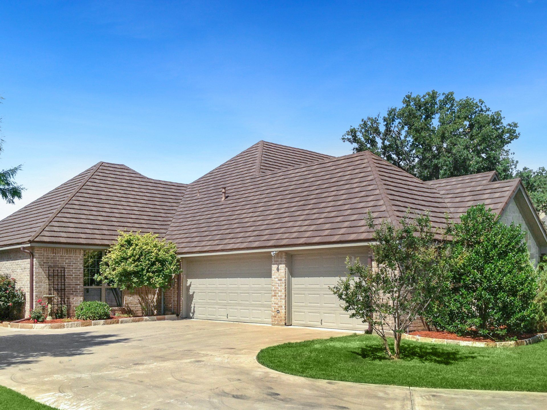 A house with a brown roof, three-car garage, and a concrete driveway under a blue sky. Green bushes and trees surround the house.