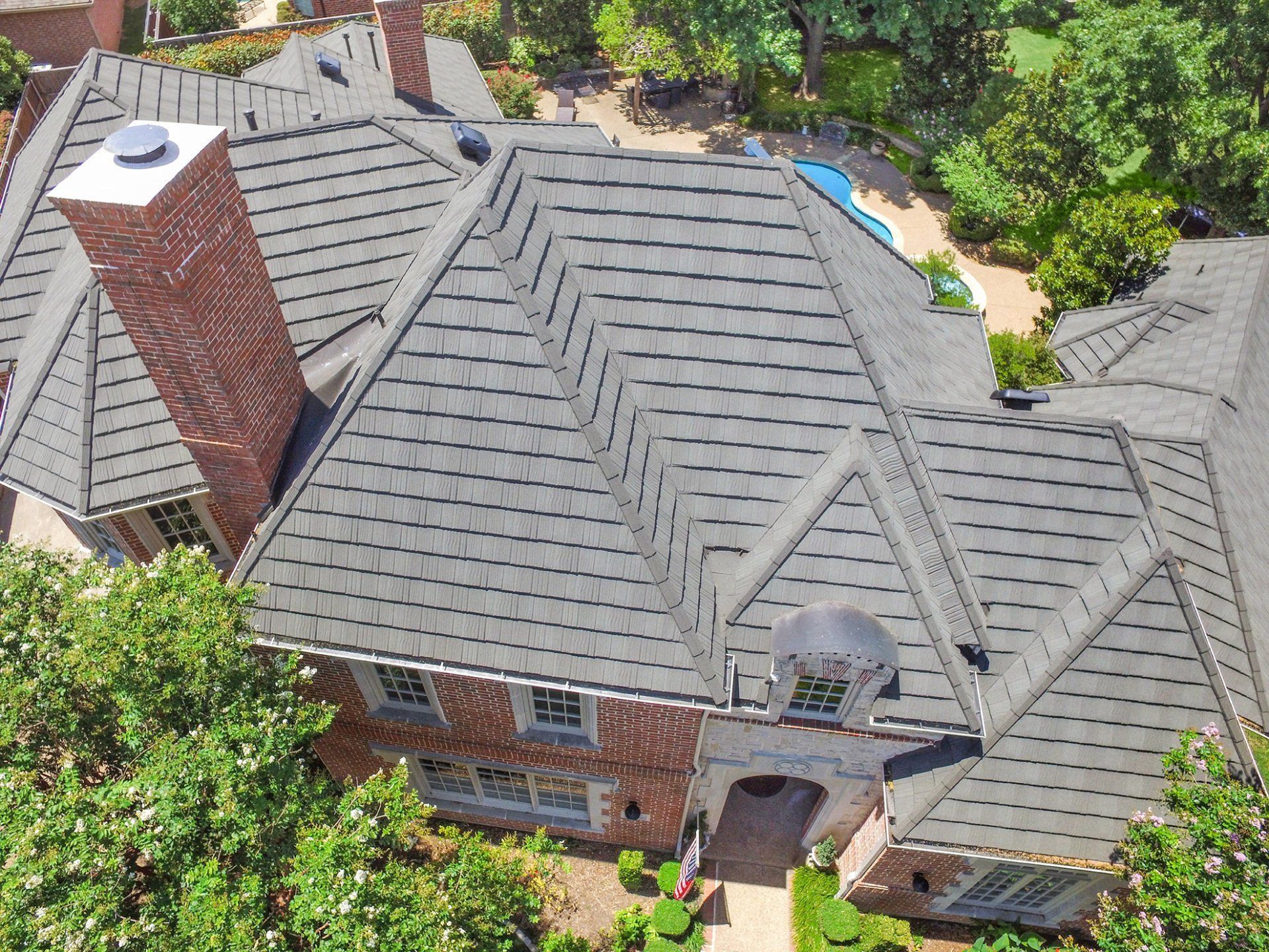 Overhead view of a large brick house with a gray roof, chimney, and surrounding trees and greenery.