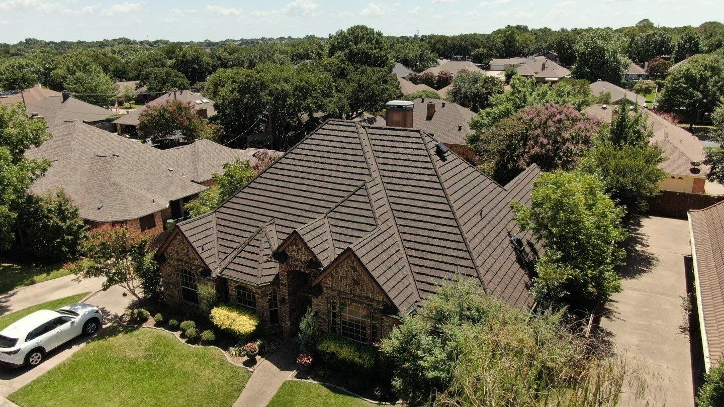 Aerial view of a large house with a brown roof in a suburban neighborhood with green lawns and trees. A white car is parked in the driveway.