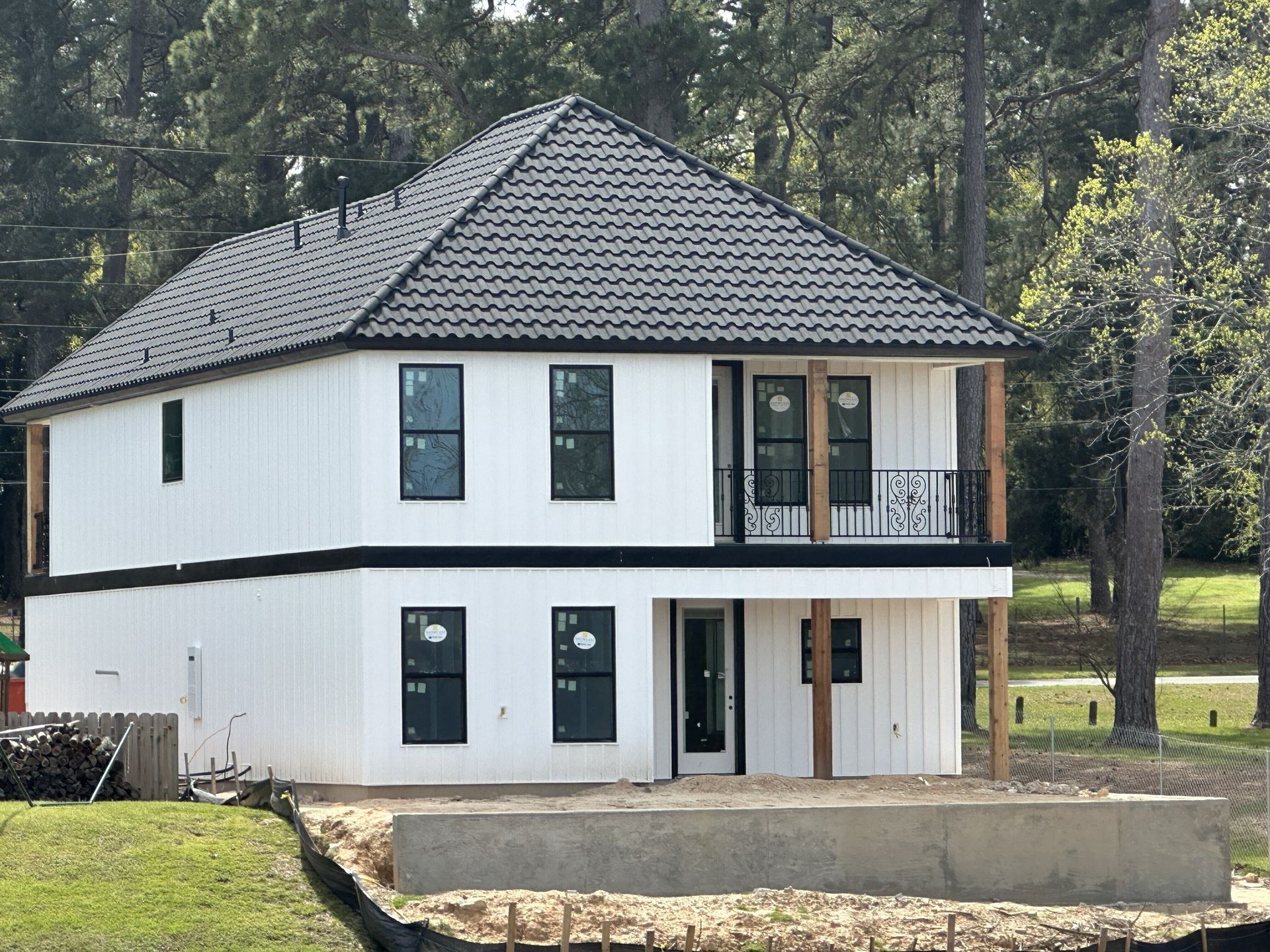 Two-story white house with a black roof and window frames under construction, with a concrete foundation and trees in the background.