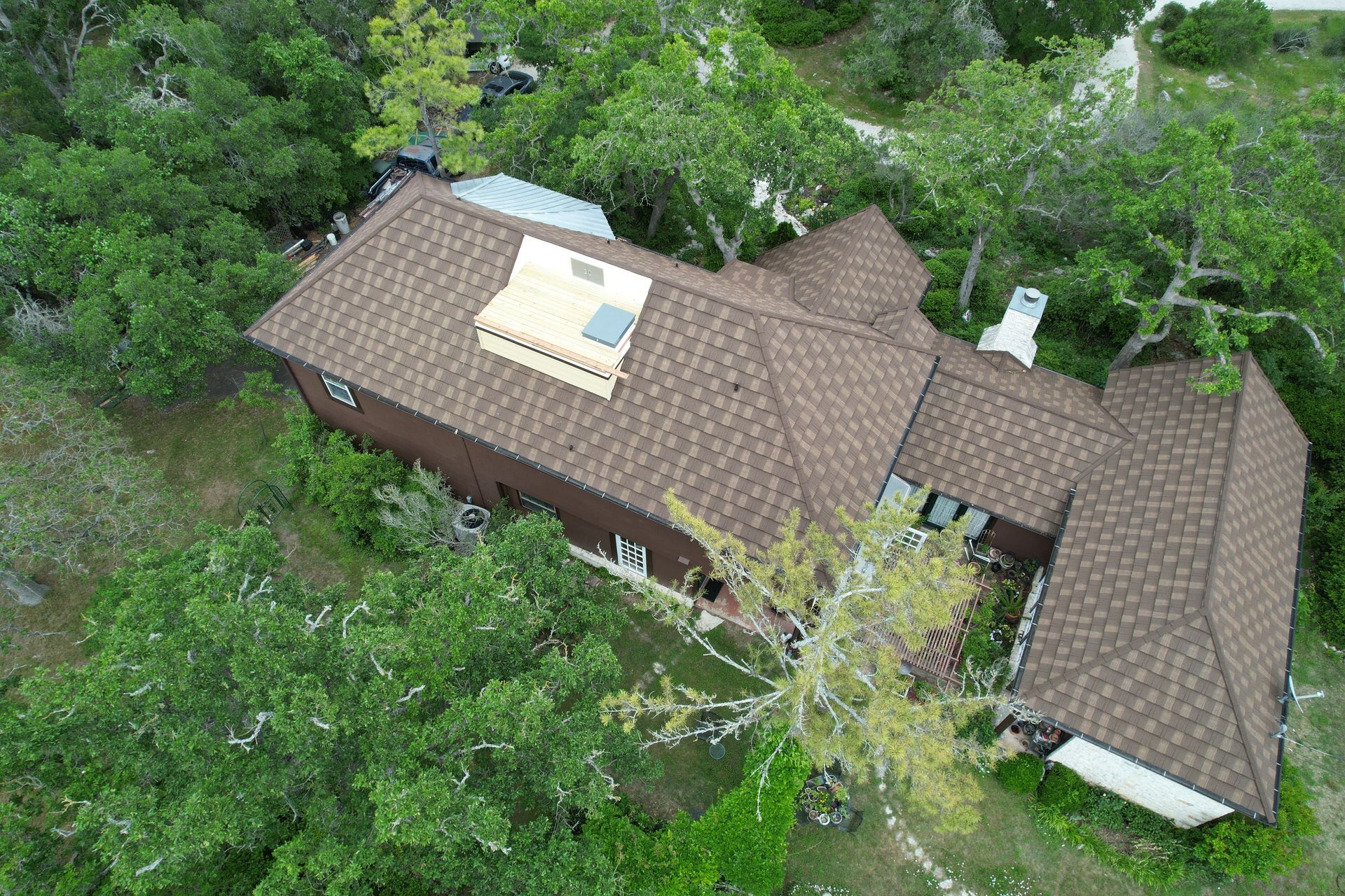 Aerial view of a brown-roofed house surrounded by green trees. A light-colored structure is visible on the roof.