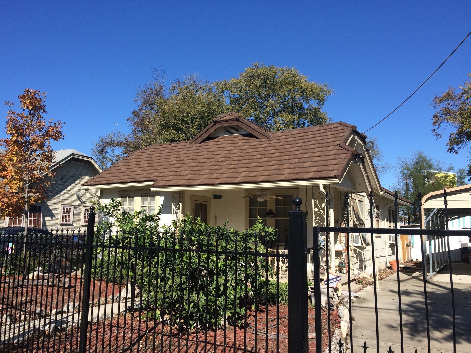Small beige house with a brown shingled roof, framed by a black fence and a clear blue sky. Bushes line the front.