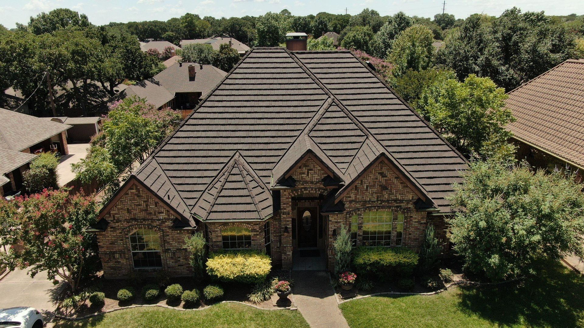 Aerial view of a brown brick house with a patterned roof and green landscaping, in a suburban setting.