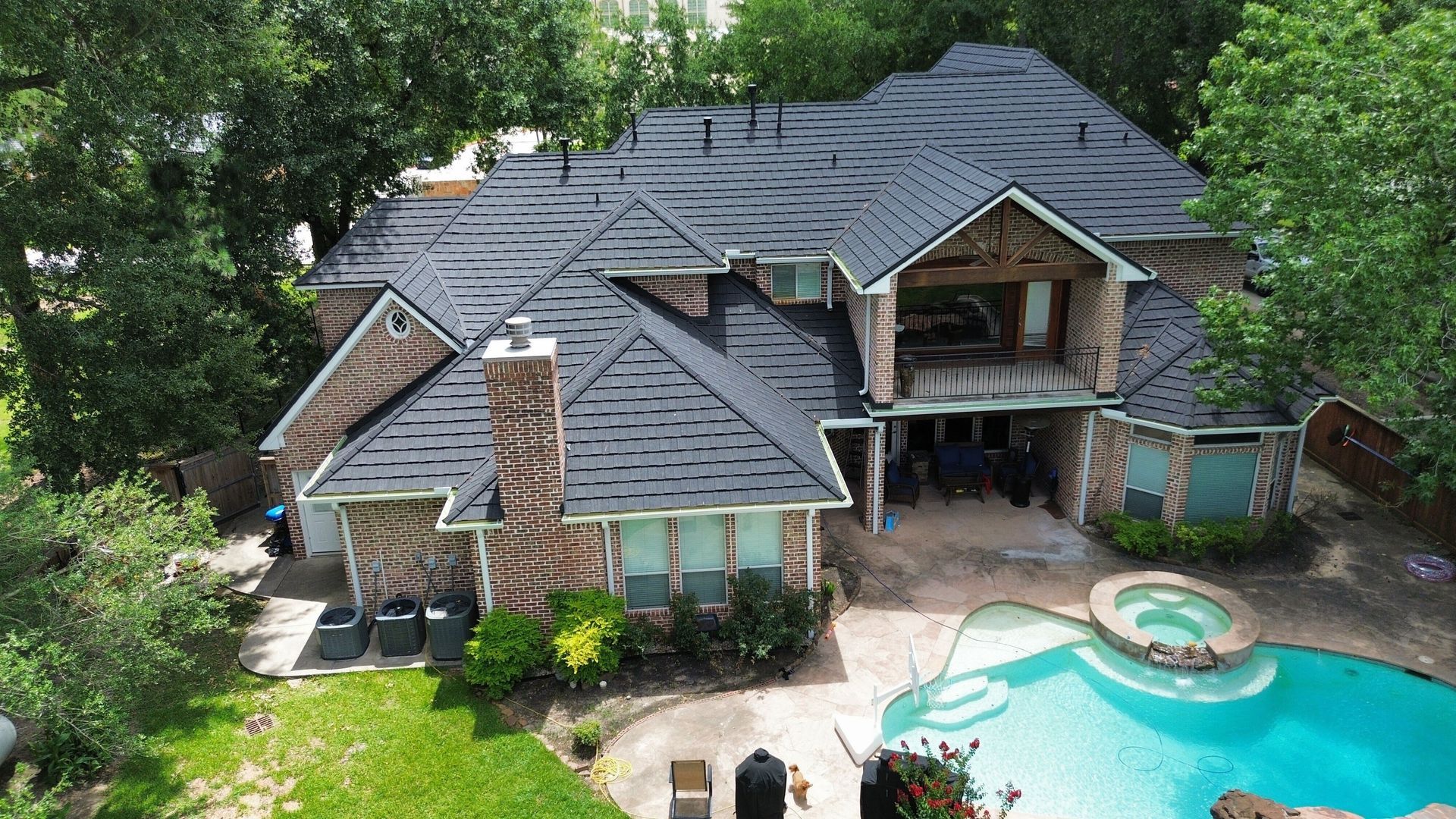 Aerial view of a two-story brick home with a dark shingle roof, a pool, and a spa.