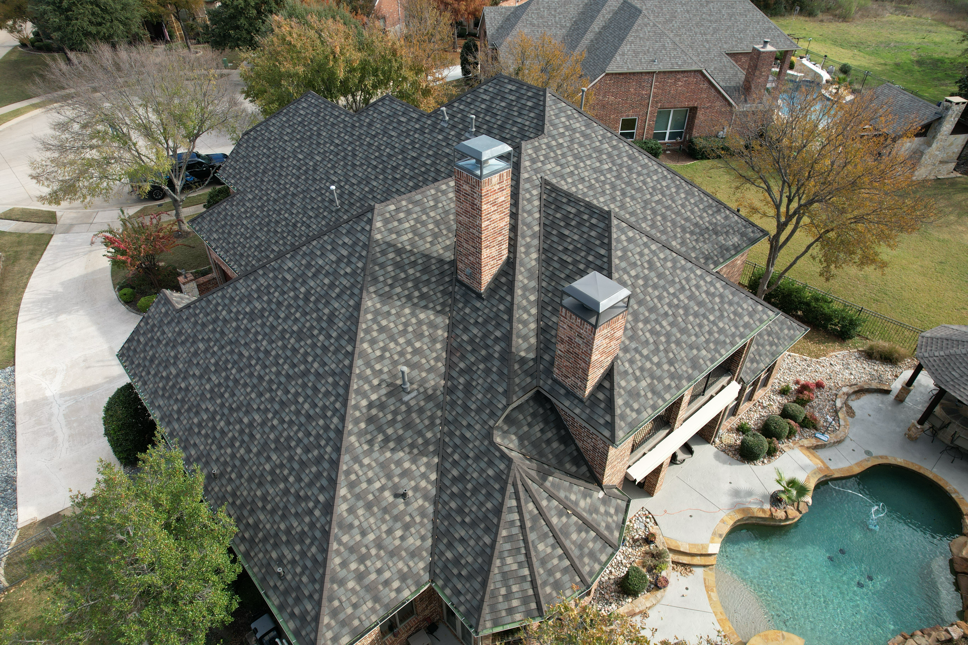 Aerial view of a house with a gray shingle roof, two brick chimneys, and a backyard with a pool.