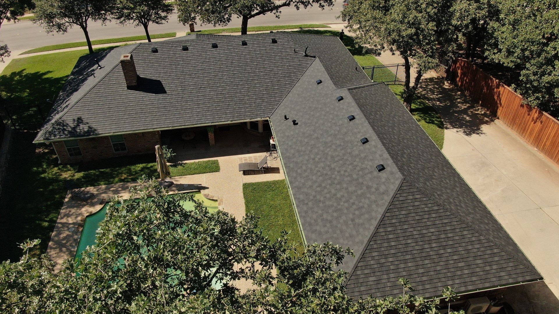 Overhead view of a one-story house with a dark gray roof, a swimming pool, and a fenced yard, in a neighborhood with trees.