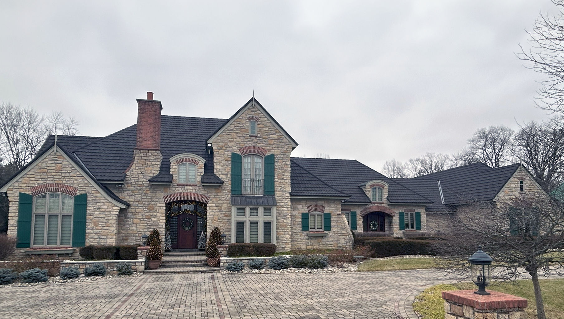 Stone house with dark roof, green shutters, and brick chimney under a cloudy sky. A brick driveway leads to the front entrance.