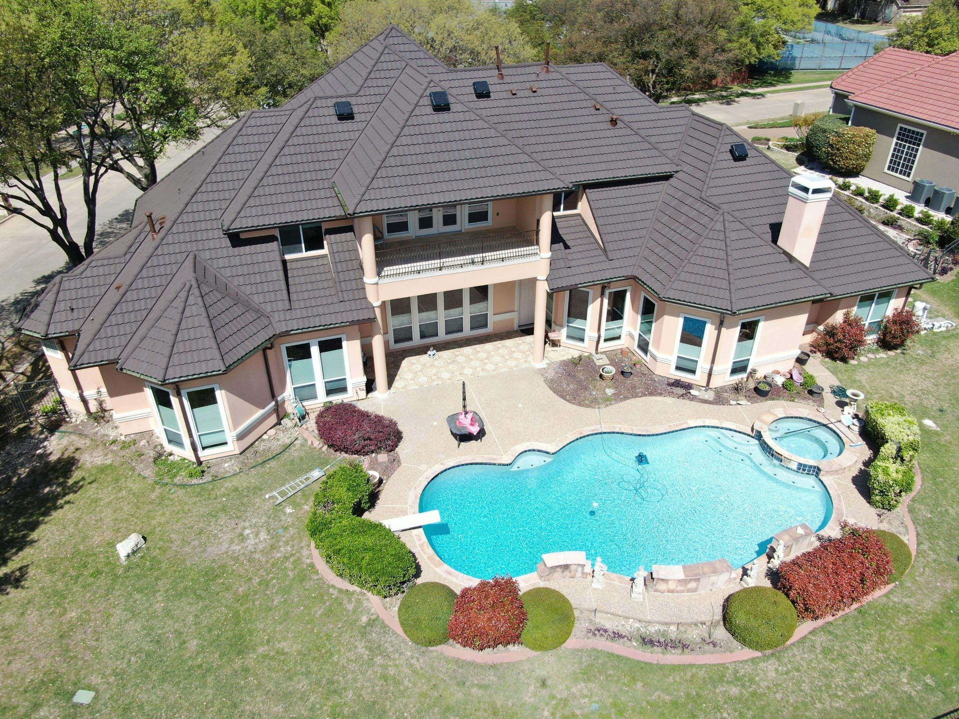 Aerial view of a large, two-story house with a dark brown roof and a pool in the backyard. The house is surrounded by greenery.