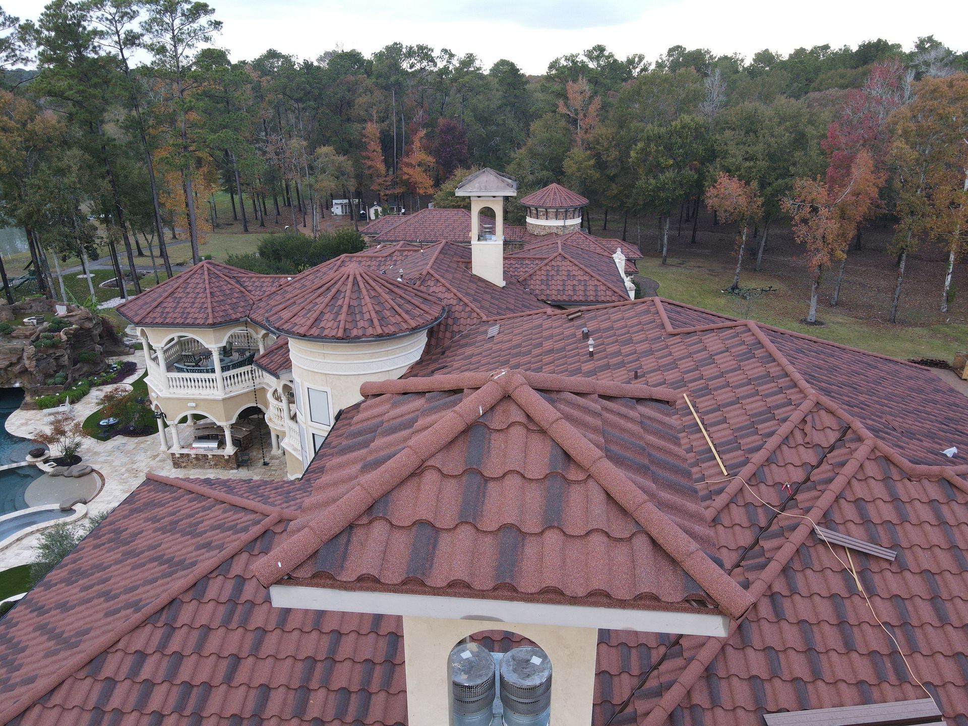 Aerial view of a large house with a red-tiled roof, surrounded by trees. A pool and landscaping are visible.