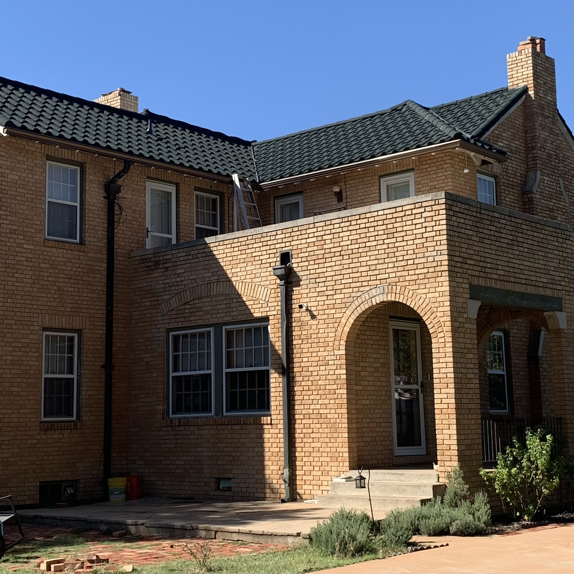 Two-story brick house with a dark green tile roof and arched entryway under a porch. Clear blue sky.
