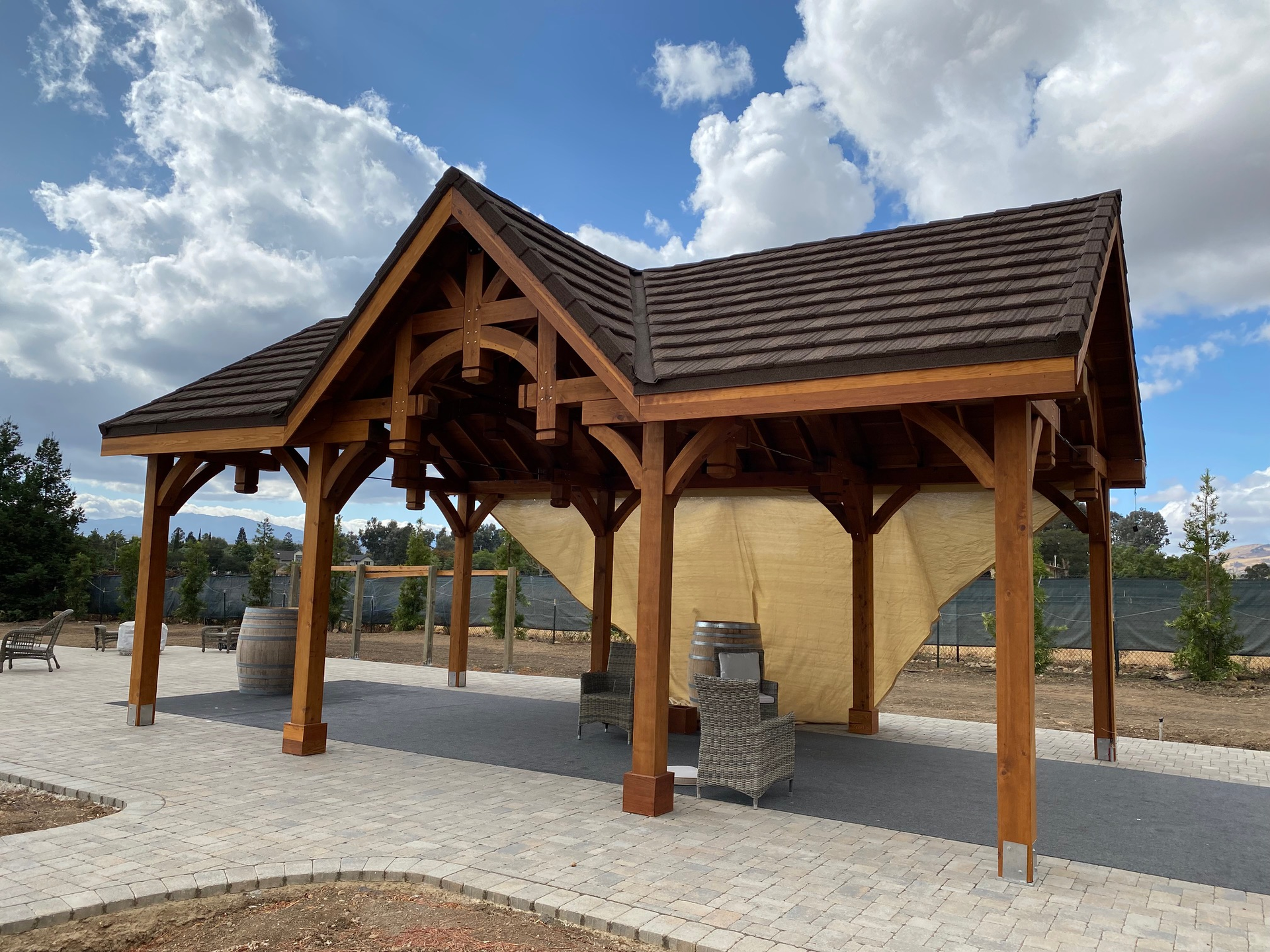 Wooden pavilion with brown roof, surrounded by a paved area and landscaping, under a partly cloudy sky.