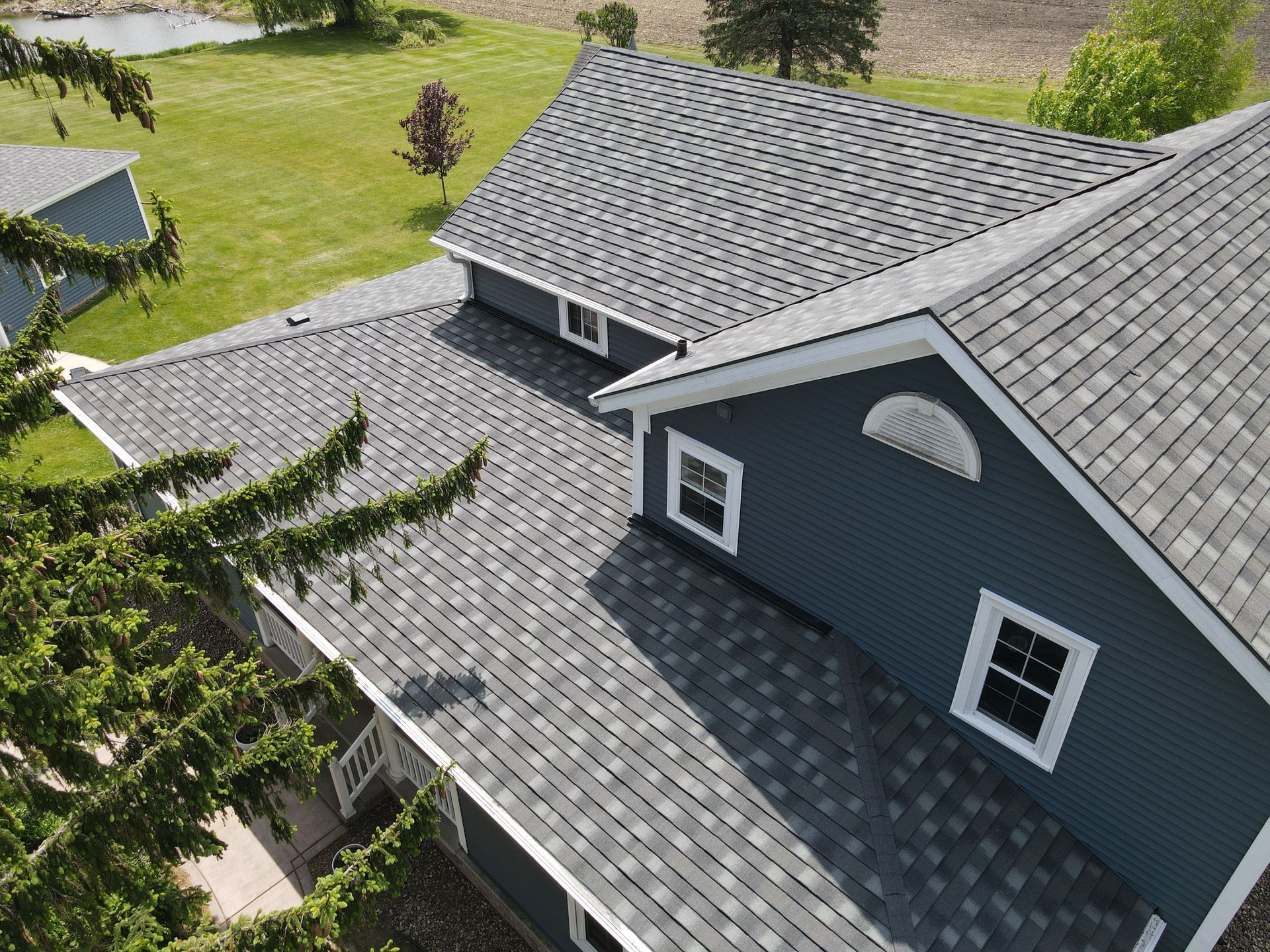 A gray shingled roof on a blue house with white trim. A shed is visible on the left and green trees surround the house.