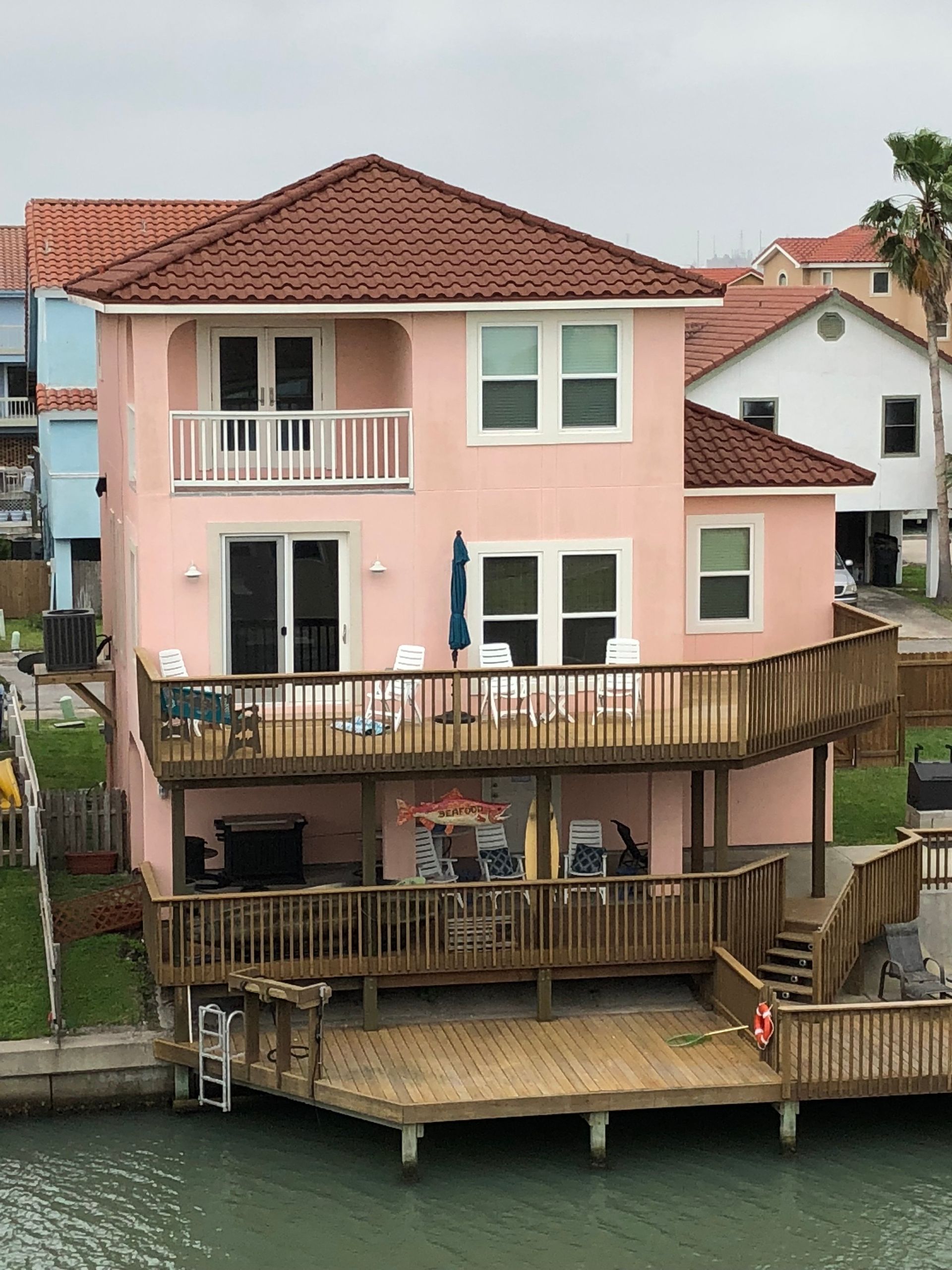 A pink stucco beach house with a red tile roof, multiple decks, and a dock on the water.