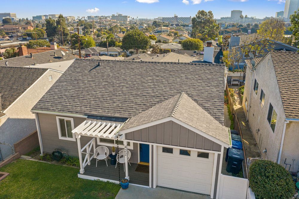 Aerial view of a house with a grey roof, a white garage door, and a blue front door. The house is in a residential neighborhood with a city skyline in the background.