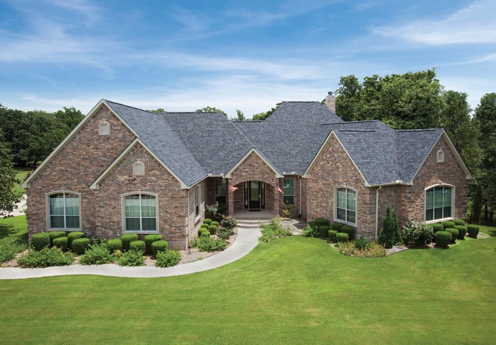 Brick house with a dark blue roof, surrounded by green lawn, trees, and shrubs under a blue sky.