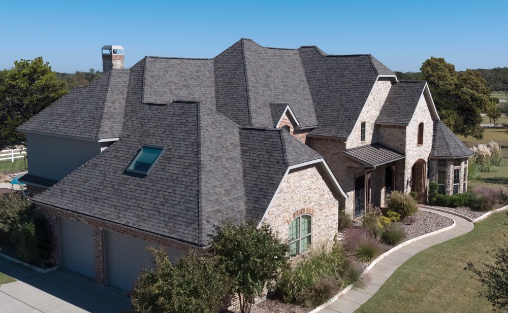 A large, multi-gabled house with gray shingles and stone facade under a clear blue sky. A walkway leads to the front door.