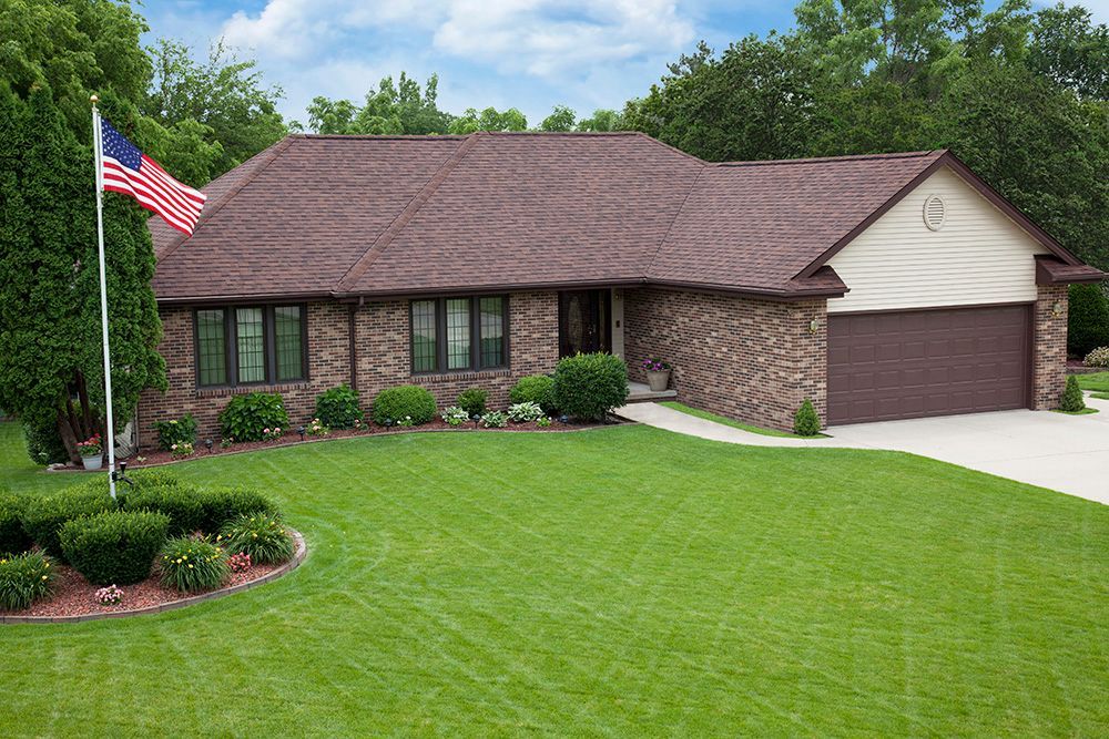 Ranch-style house with brown brick exterior, brown roof, and a green lawn; an American flag waves in the front yard.