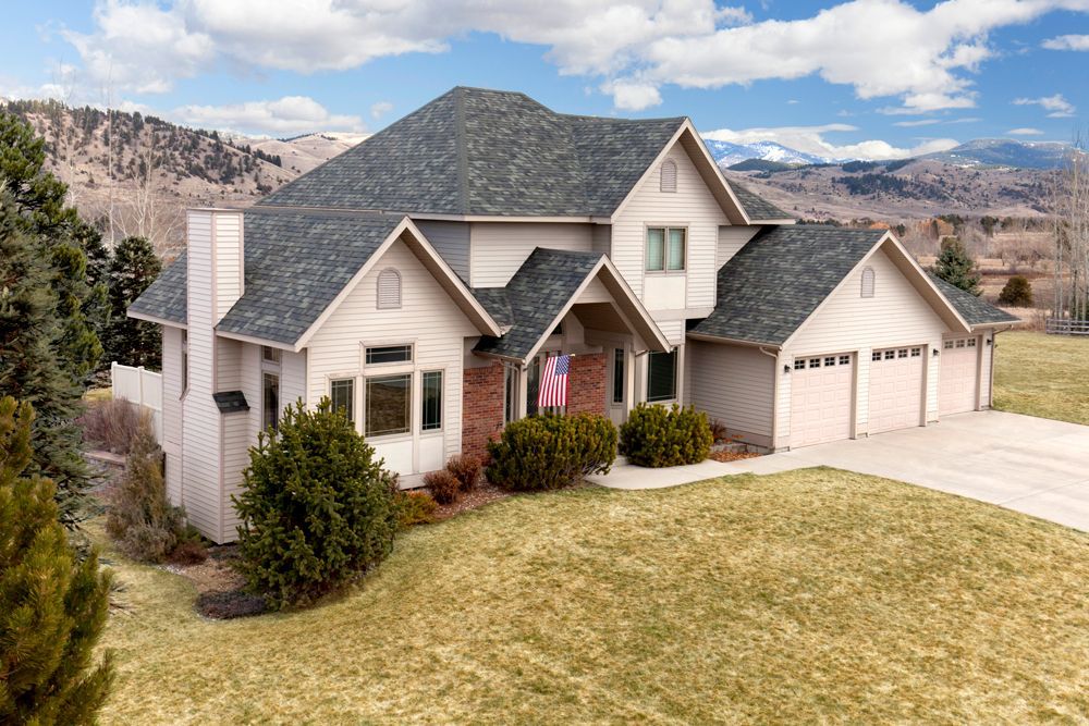 Two-story beige house with a gray roof, three-car garage, and American flag, set against a backdrop of mountains.