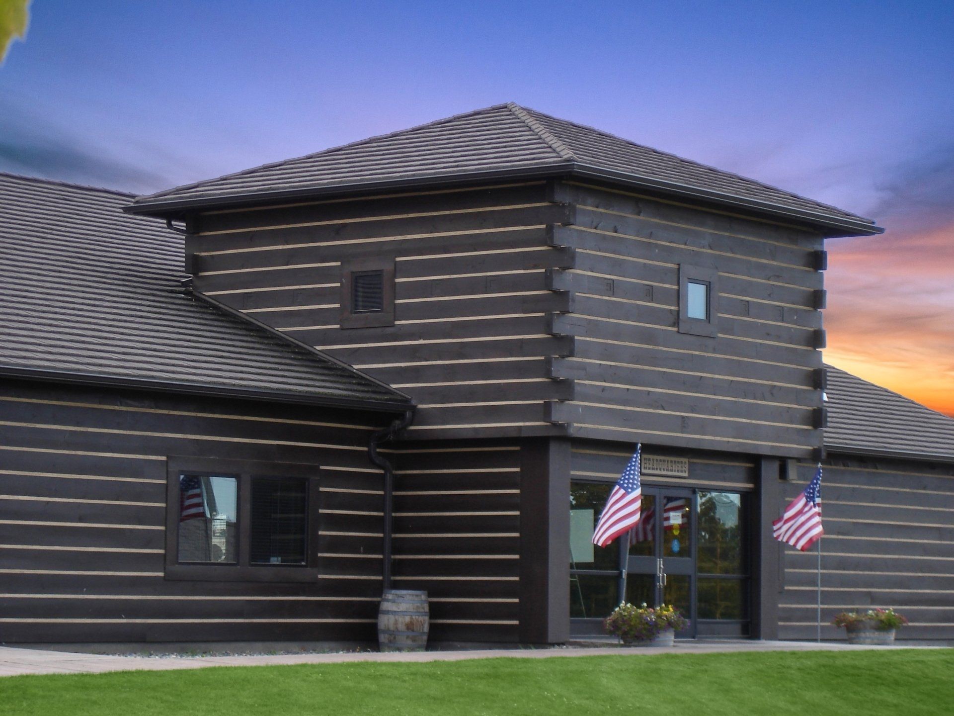 A building with dark log siding and a square tower entrance. American flags flank the door under a colorful sunset sky.