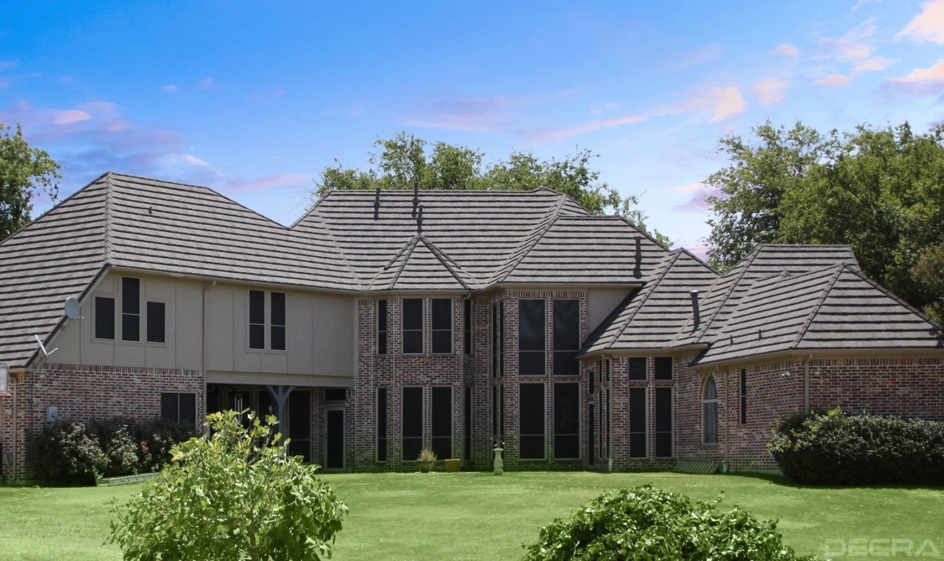 Large brick and beige house with a tiled roof, green lawn, and trees under a blue sky.
