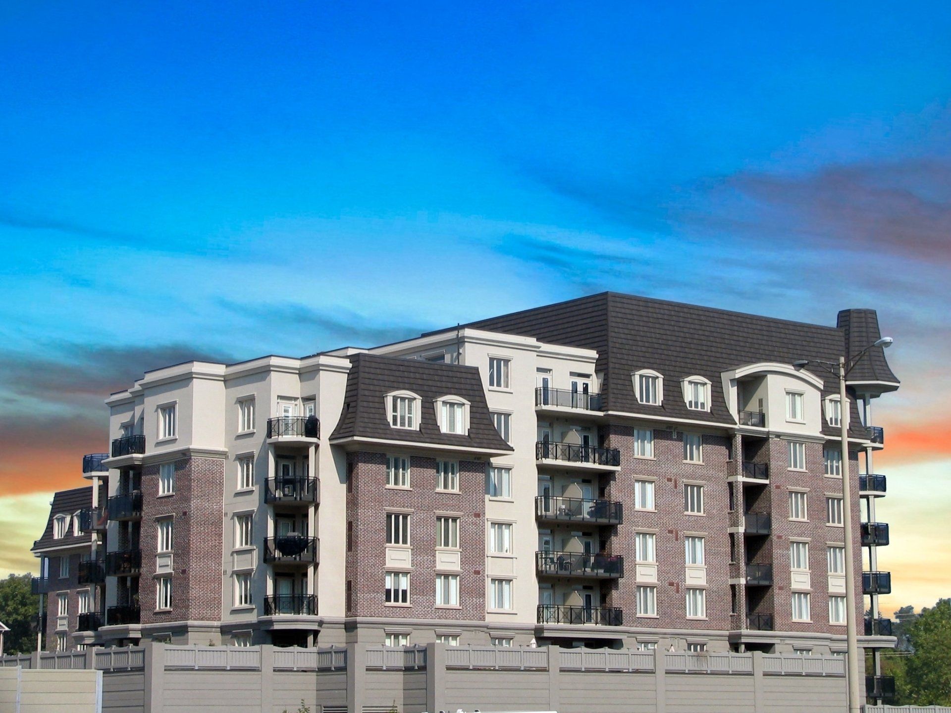 Multi-story brick and beige apartment building with black roof against a blue and orange sky.