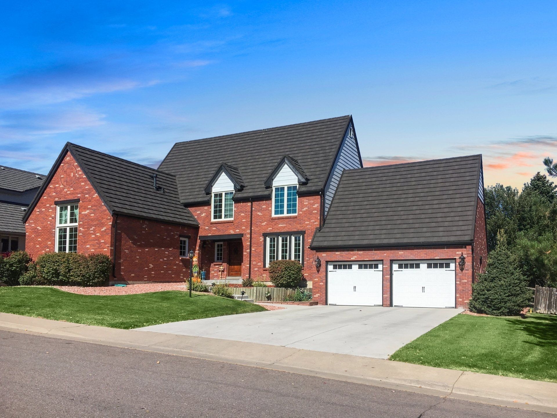 Red brick house with black roof, two-car garage, and driveway against a blue sky.