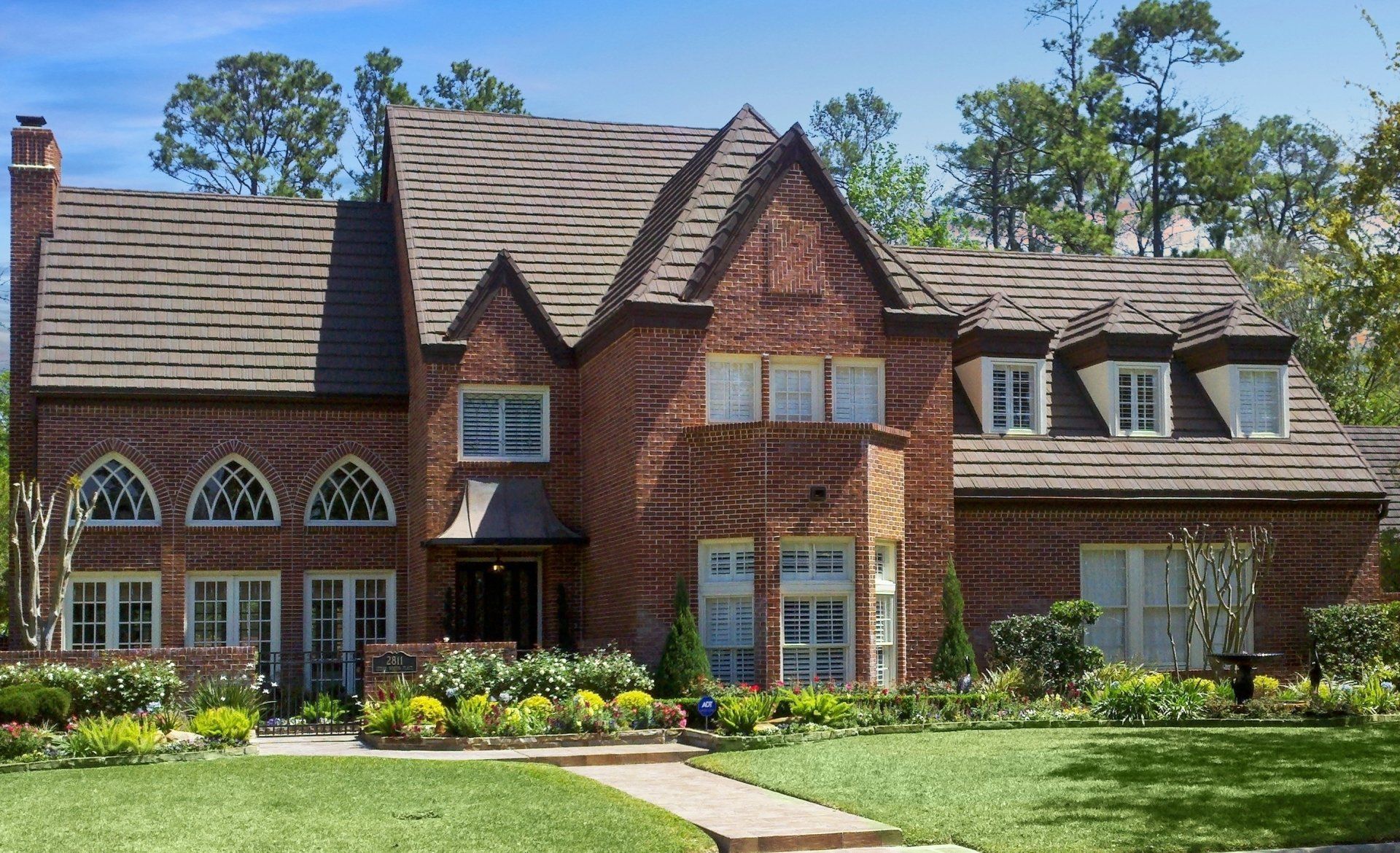 Two-story brick Tudor-style home with a manicured lawn and garden on a sunny day.