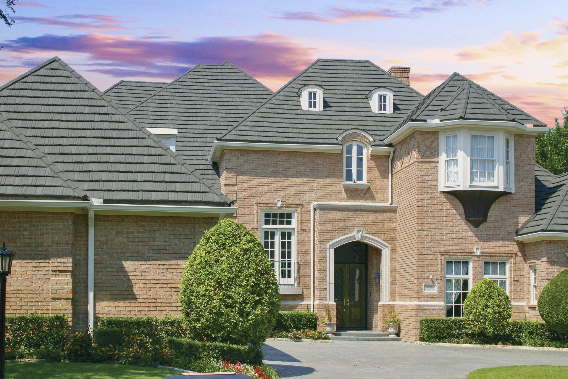 Large brick house with gray shingled roof, manicured bushes, and bay window under a cloudy sky.