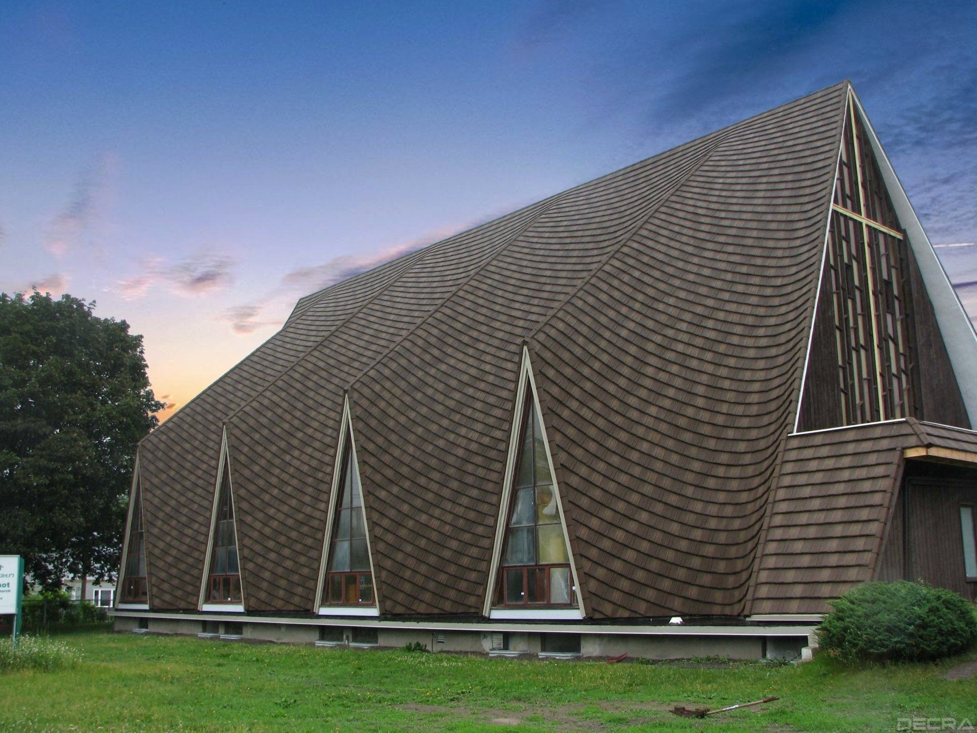 Church with a brown, curved roof and triangular windows. A cross is visible on the side. Set on a lawn under a twilight sky.