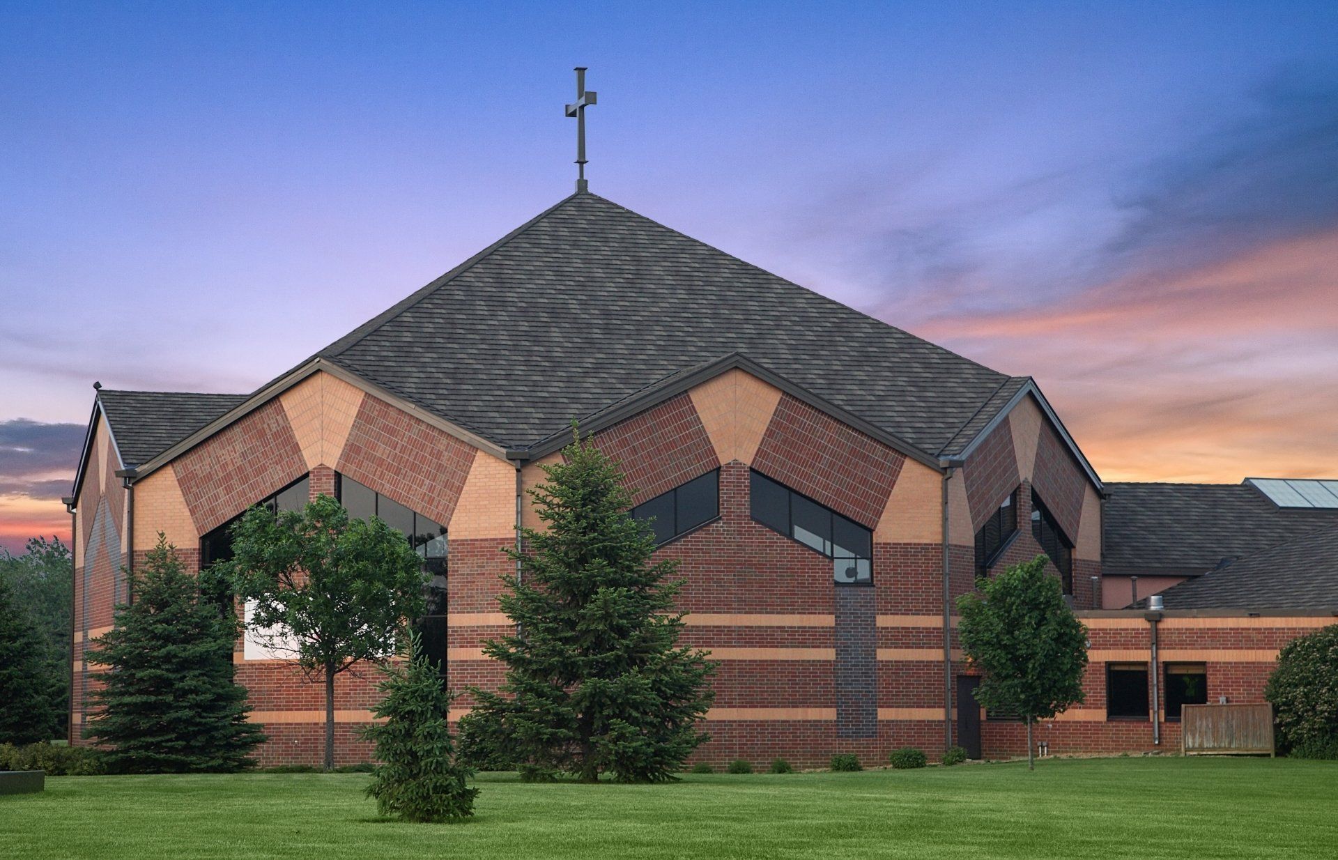 Church with red brick facade, gabled roof, and cross on top, set against a dusk sky. Green grass and trees in the foreground.