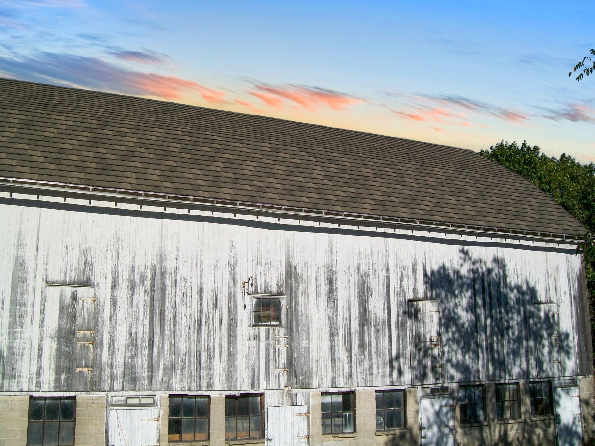 Weather-beaten white barn with a gray roof against a colorful sunset sky. Shadow of a tree on the barn.