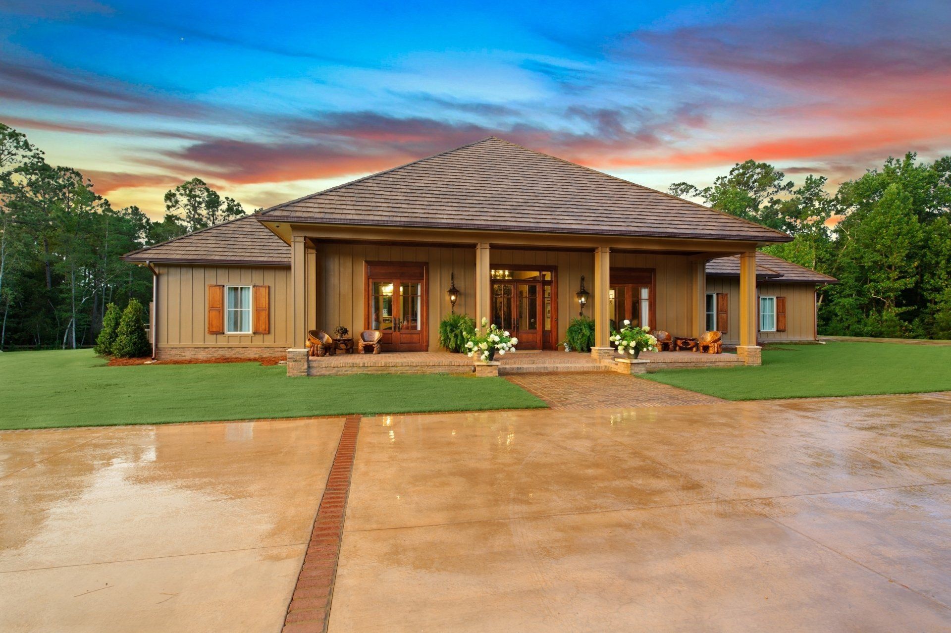 A large, tan-colored house with a covered porch and columns, set against a sunset sky and surrounded by green grass and trees.