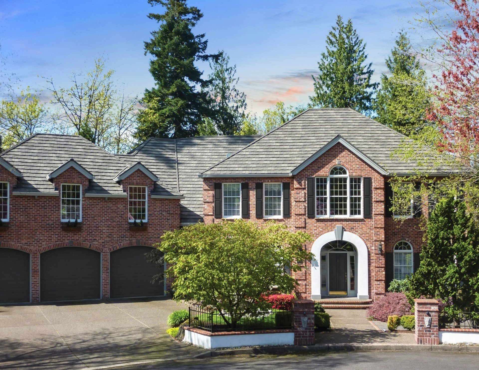 Brick two-story house with a three-car garage, gray roof, and arched entryway. Trees and bushes frame the front.