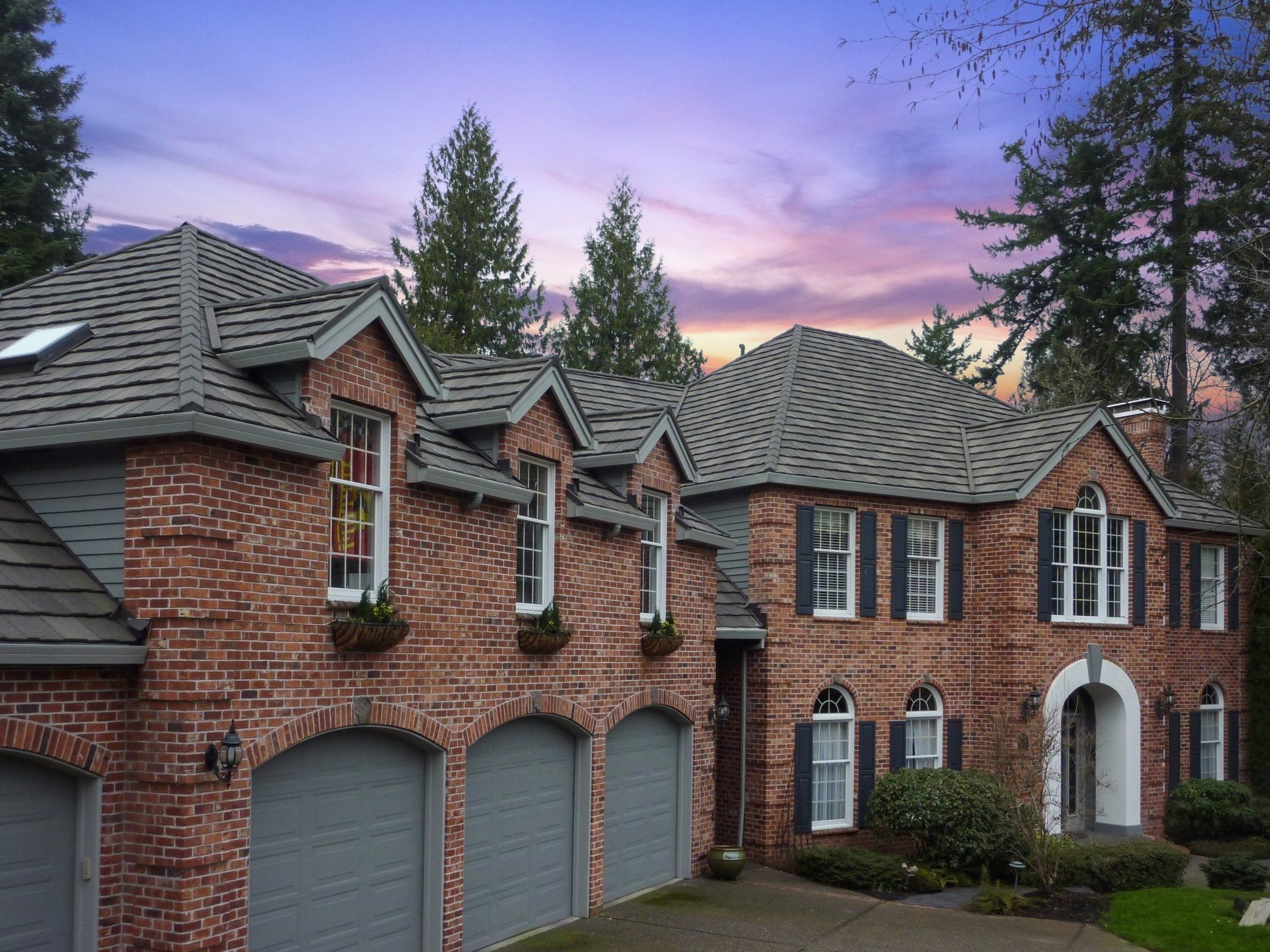 Brick home with a gray roof, multiple windows, and garage doors.  The sky is a colorful blend of pinks, purples, and blues.
