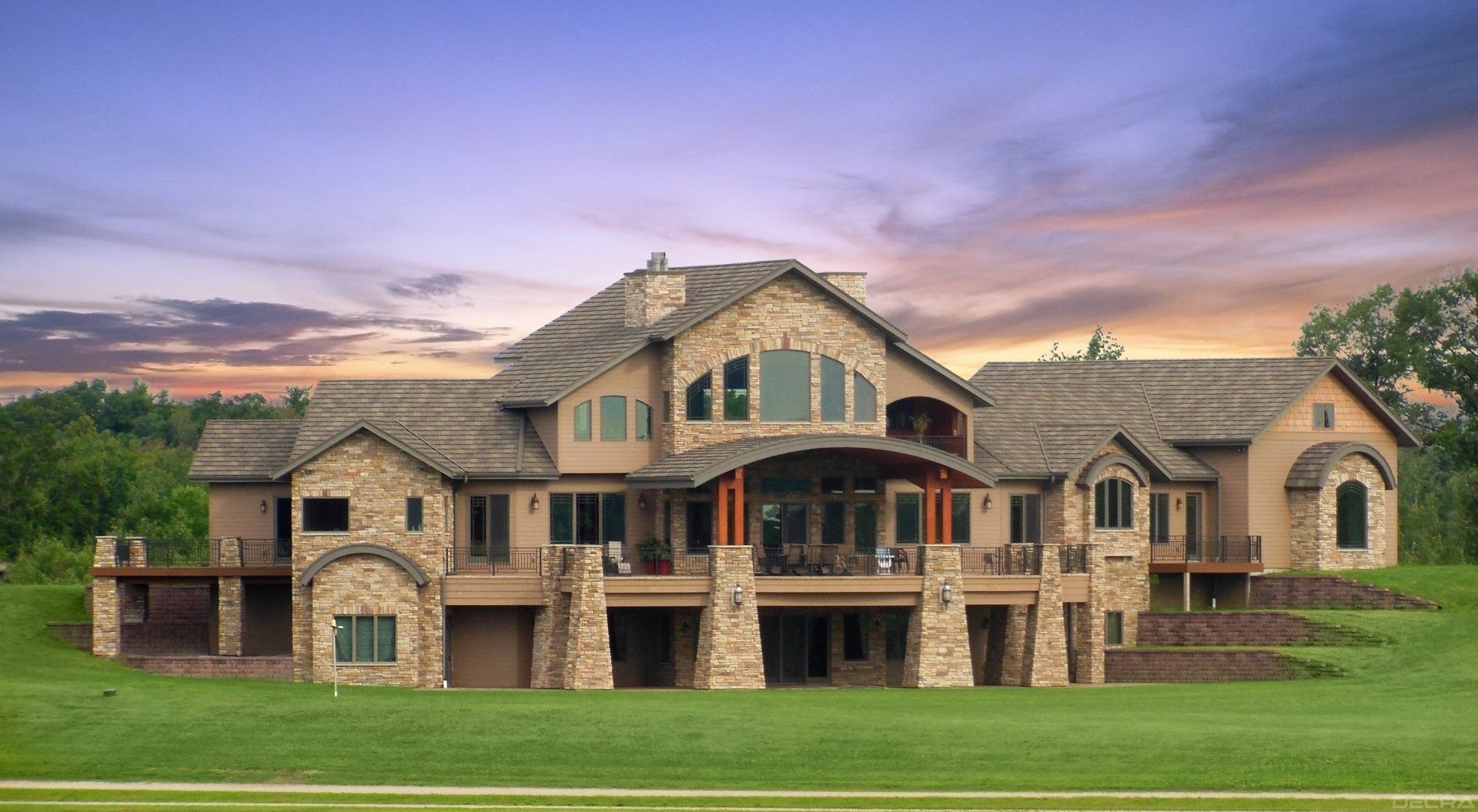 Large stone mansion on a green lawn under a dusky sky. Features multiple levels, balconies, and a covered patio.