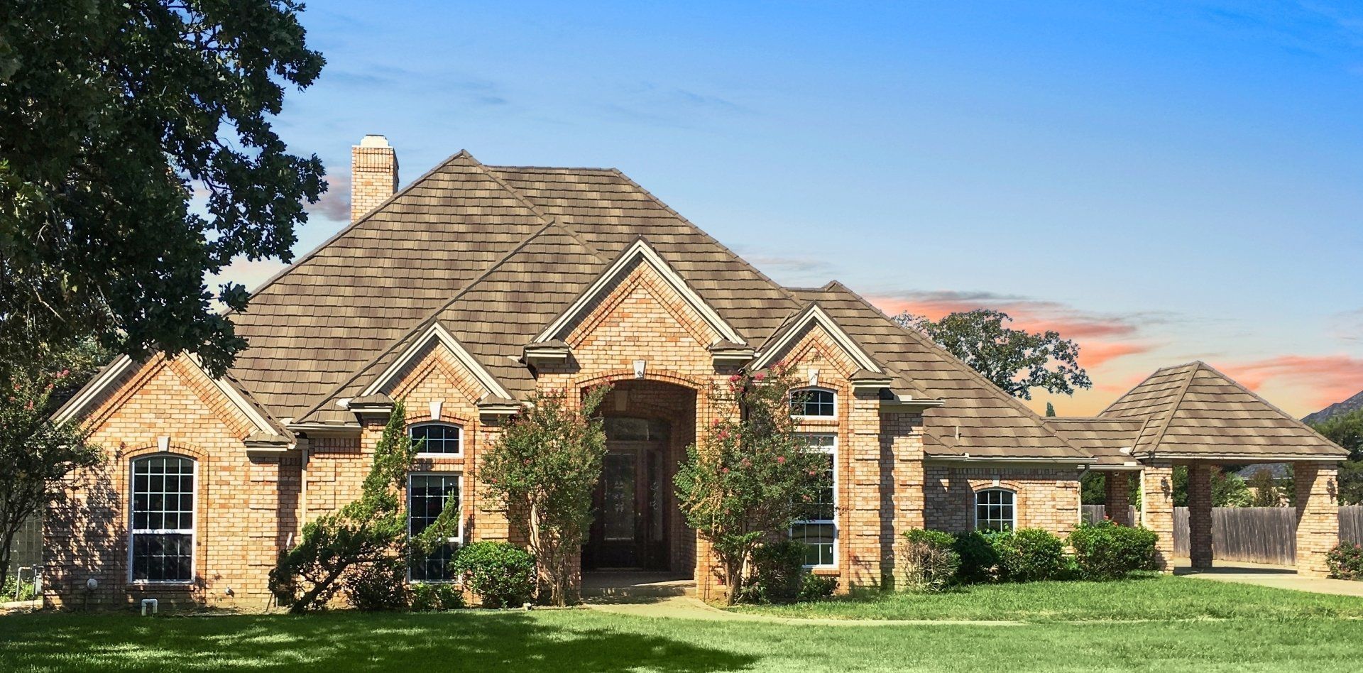 A large brick house with a brown shingle roof. The house has a green lawn and is set against a blue sky.