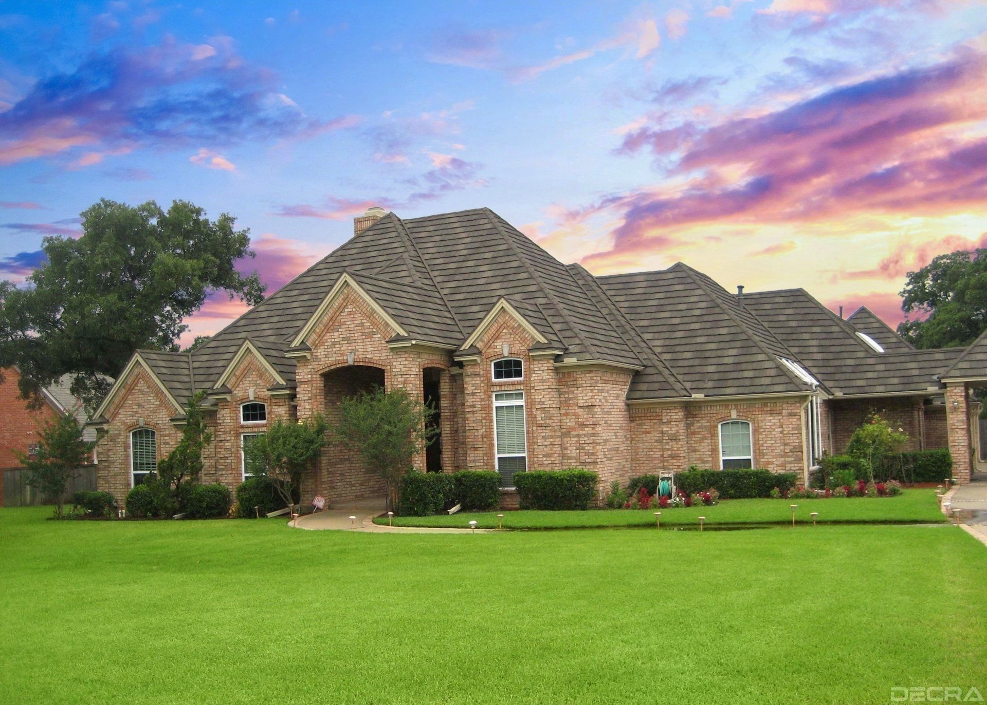 Brick house with a brown shingled roof on a grassy lawn under a vibrant, colorful sky.