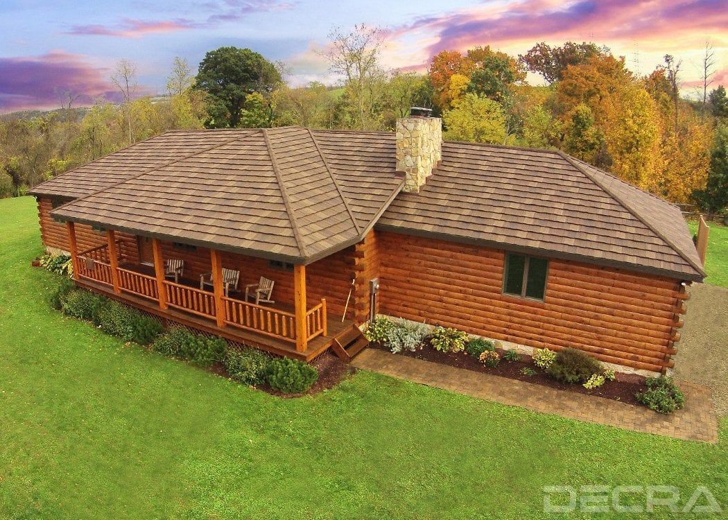 Rustic log cabin with a wraparound porch and brown shingle roof. Set in a grassy area with fall foliage in the background under a colorful sunset.
