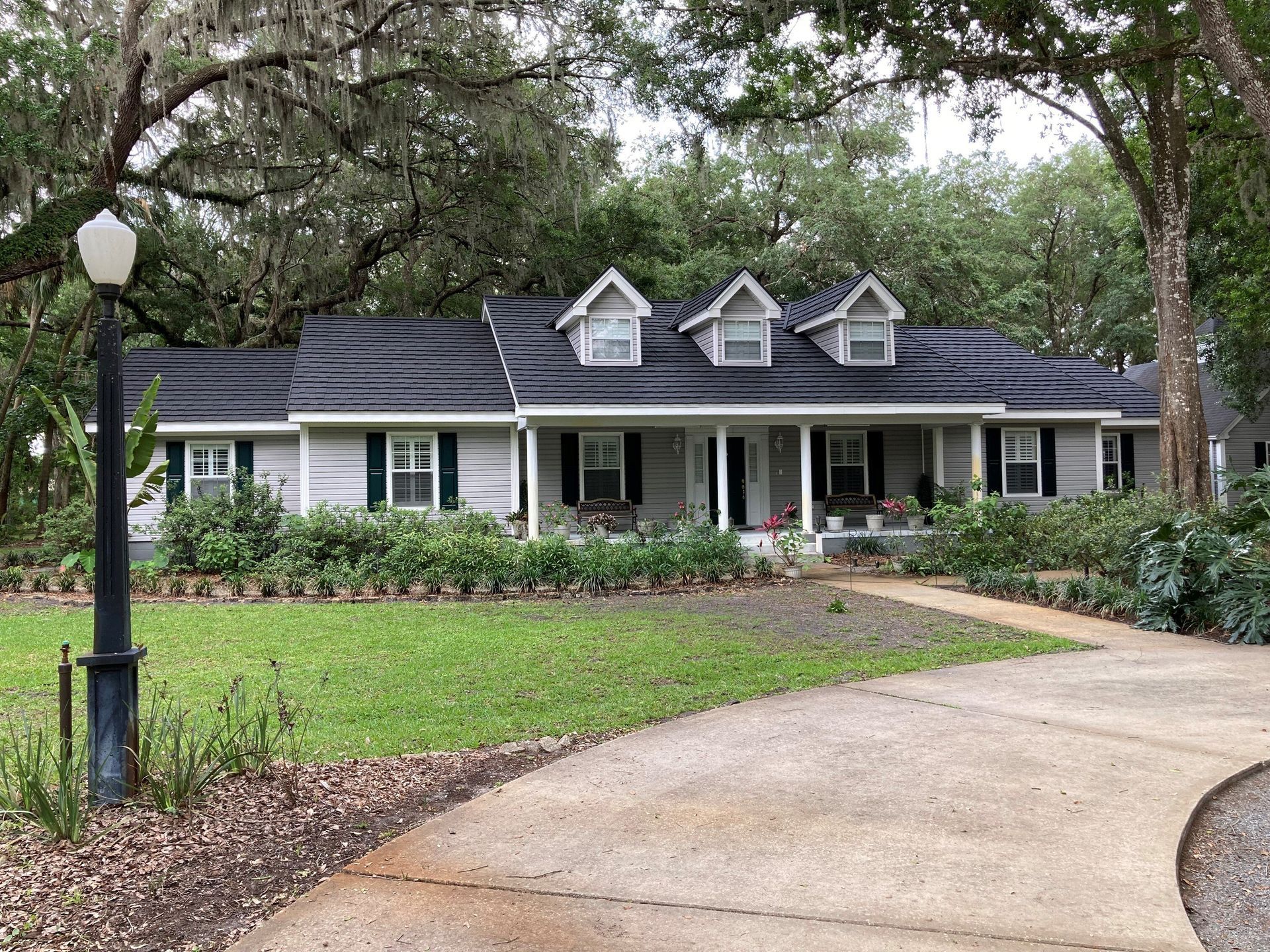 A light gray ranch-style house with a black roof, three dormers, and a curving driveway in a lush, green setting.