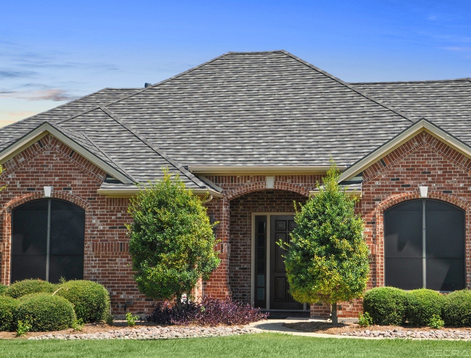 Brick house with a gray roof, arched doorway, and green shrubbery against a blue sky.
