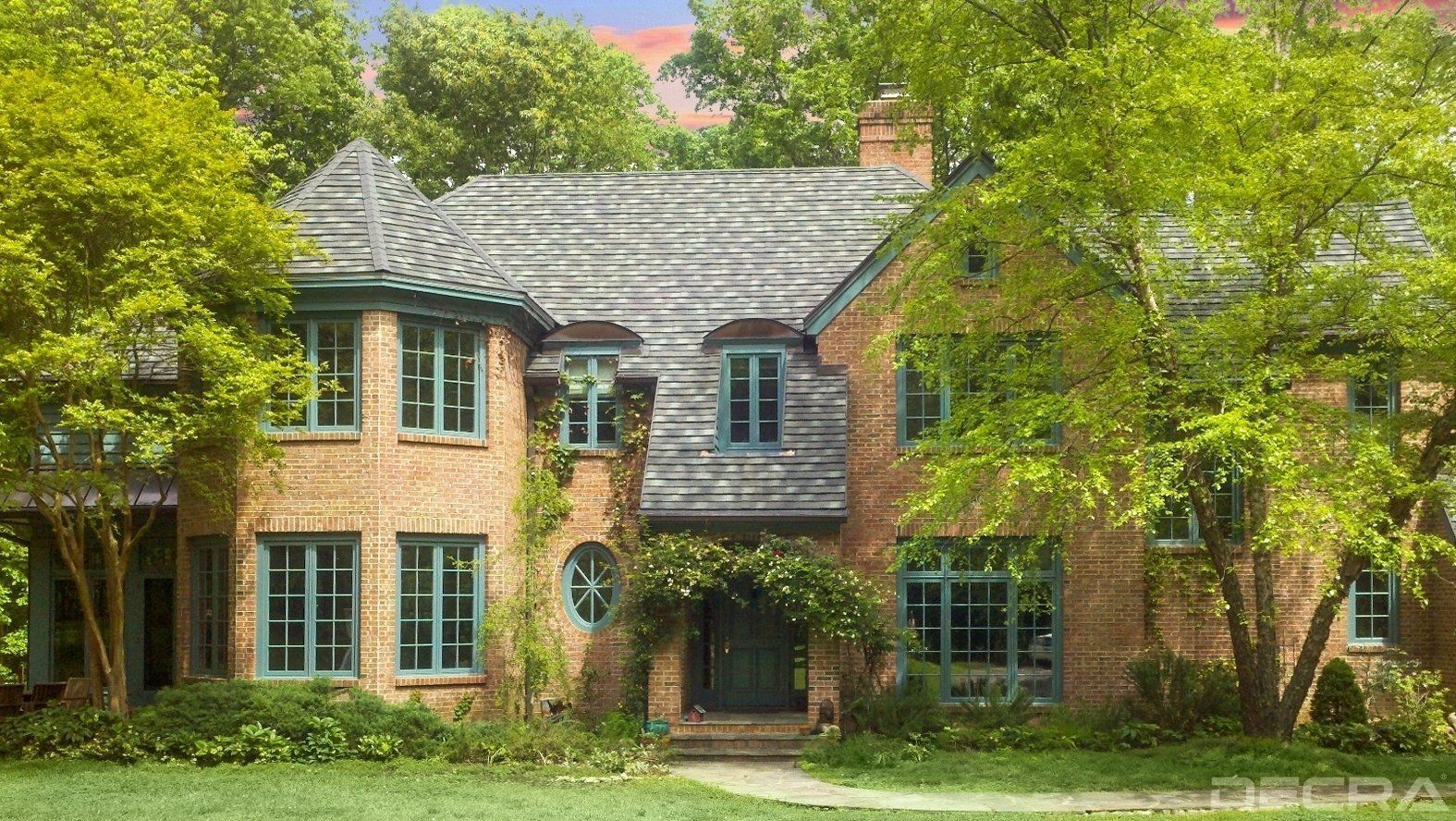 Brick house with green-trimmed windows and a gray shingled roof. Lush green trees surround the building.
