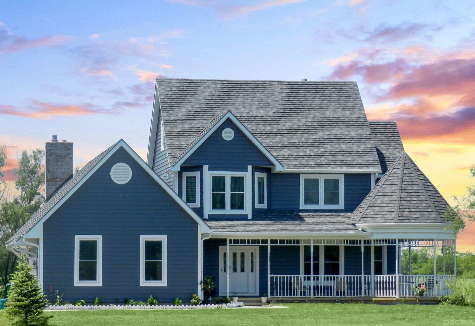 Blue two-story house with white trim and porch, set against a sunset sky and grassy lawn.