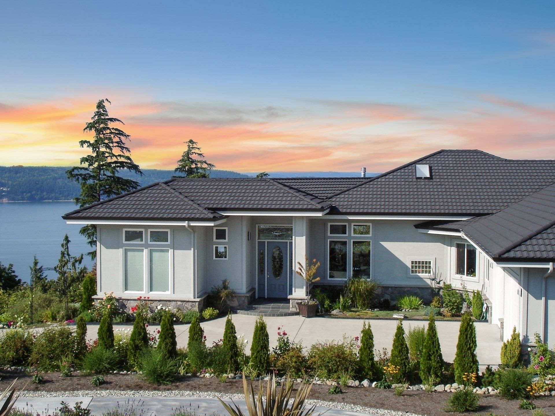 A light-colored house with a dark gray roof sits on a hill overlooking a body of water at sunset.