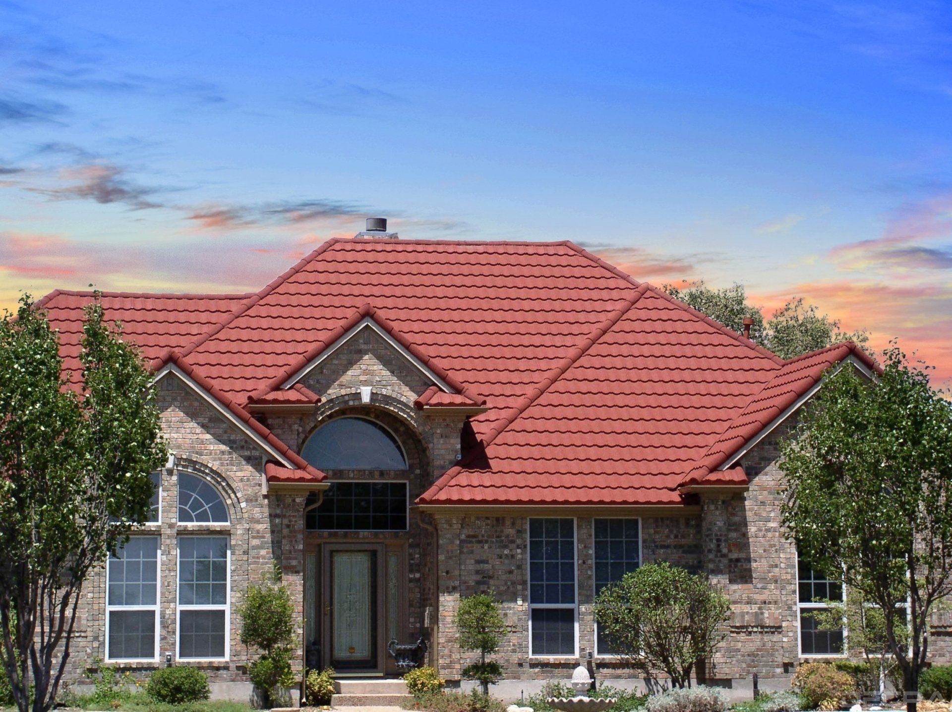 Brick house with red tile roof under a blue and orange sky. Front door and multiple windows with small trees in front.