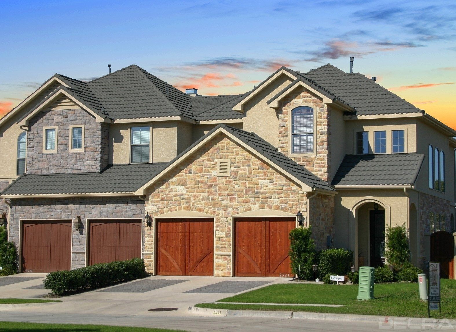 Two-story house with stone and tan exterior, brown garage doors, and a green lawn.