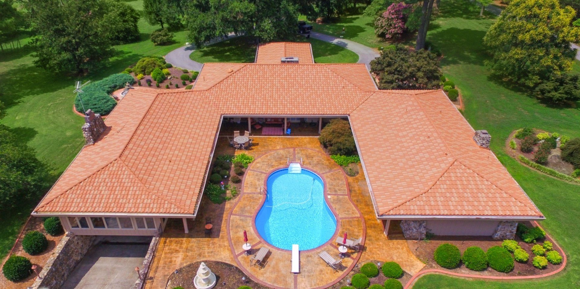 Aerial view of a large U-shaped house with a blue pool in the center. The house has a brown roof, surrounded by green grass and trees.